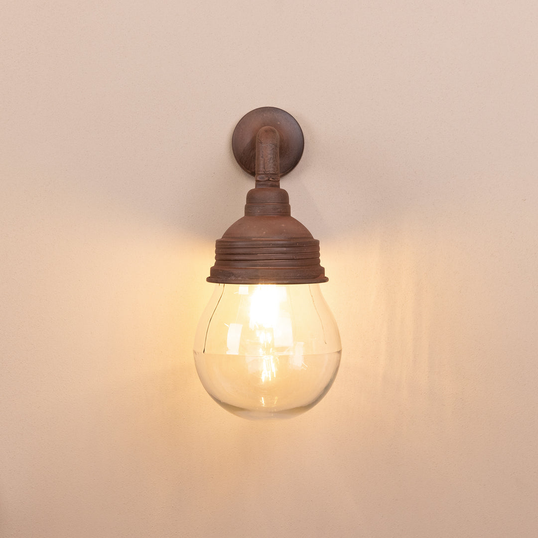 Front on image of the Agriframes Trowbridge Outdoor Wall Lamp in a rust finish with its bulb illuminated, mounted on a neutral background. 