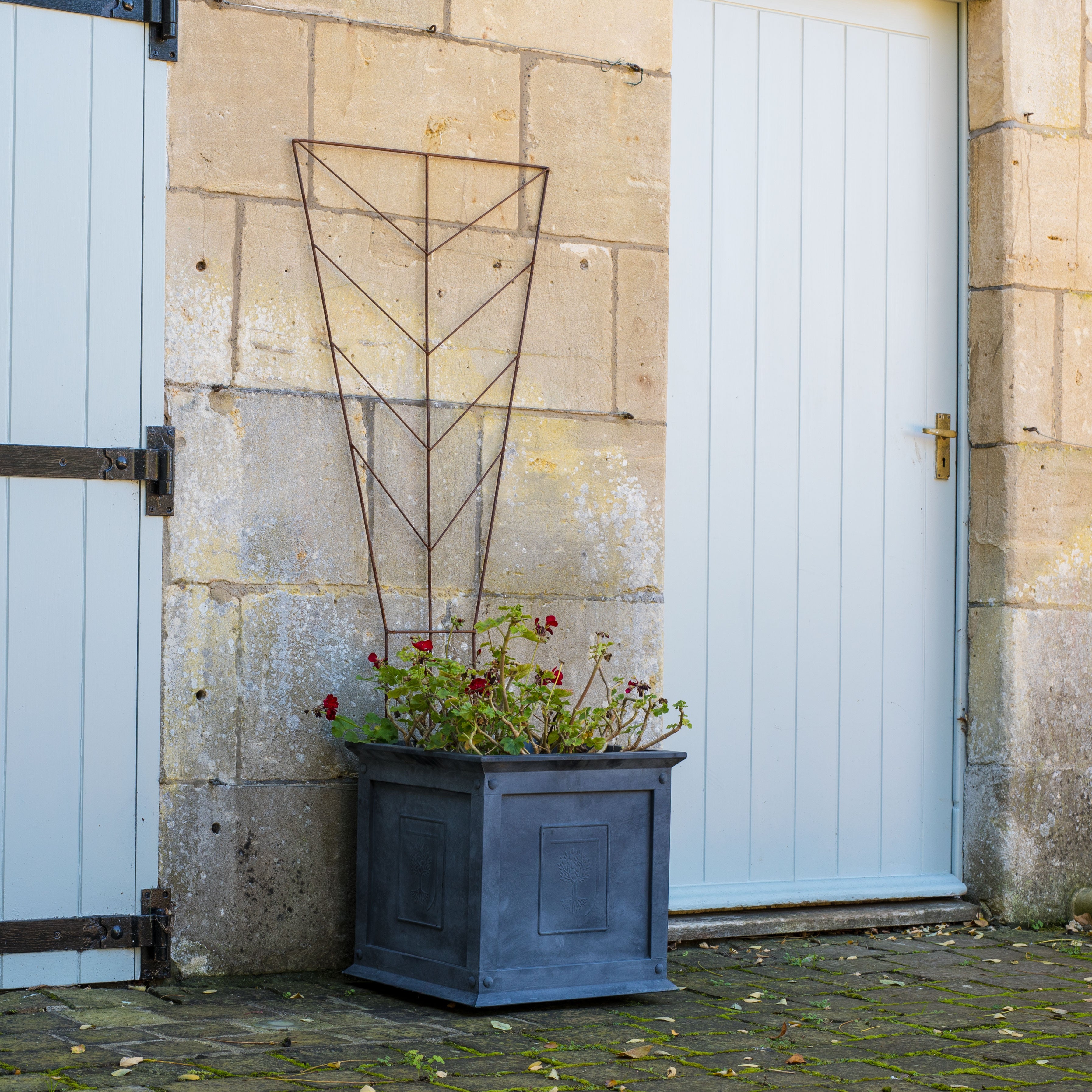Angled view of a rust-coloured Agriframes deco metal trellis with plants in an Agriframes metal zinc planter against a stone wall.