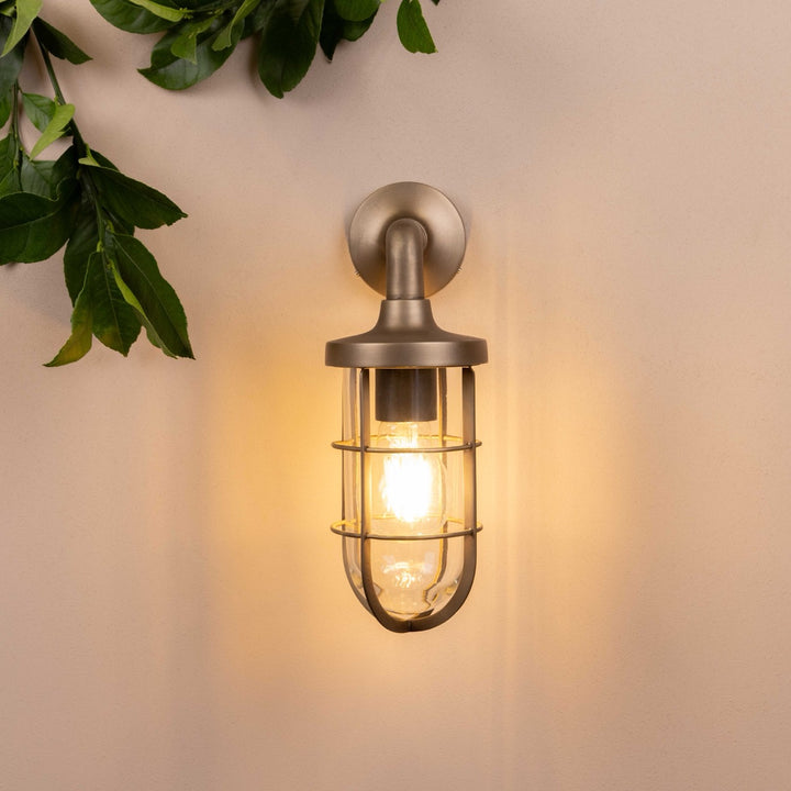 Front on image of the Agriframes Exmouth Outdoor Wall Lamp in a Brass finish with its bulb illuminated, mounted on a neutral background. 