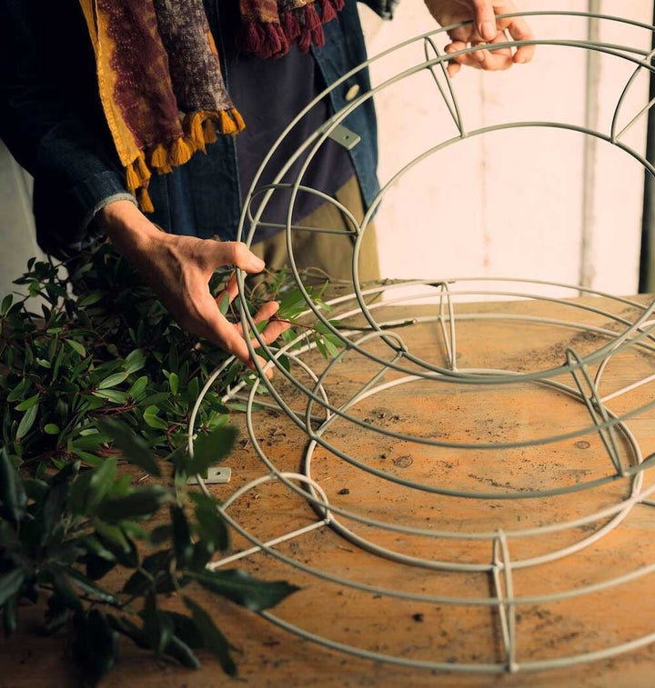 Two agriframes florist wreath trellis in a sage green finish on a wooden table, with a woman handling one of the trellis.