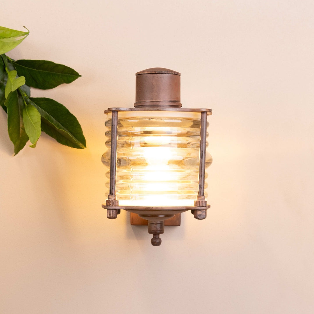 Front on image of the Agriframes Frome Outdoor Wall Lamp in a Rust finish with its bulb illuminated, mounted on a neutral-coloured wall.