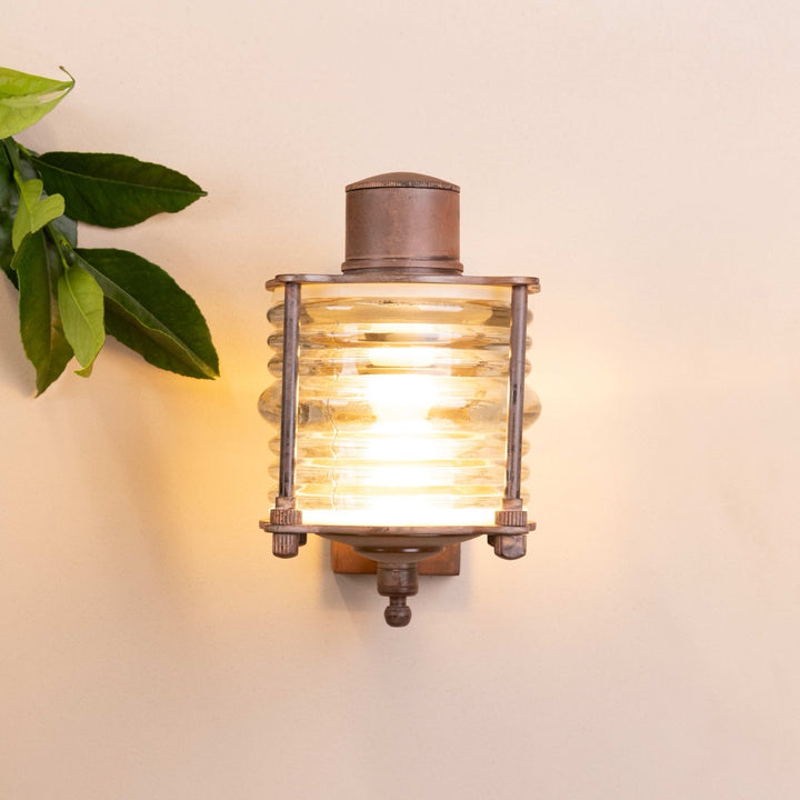 Front on image of the Agriframes Frome Outdoor Wall Lamp in a Rust finish with its bulb illuminated, mounted on a neutral-coloured wall.
