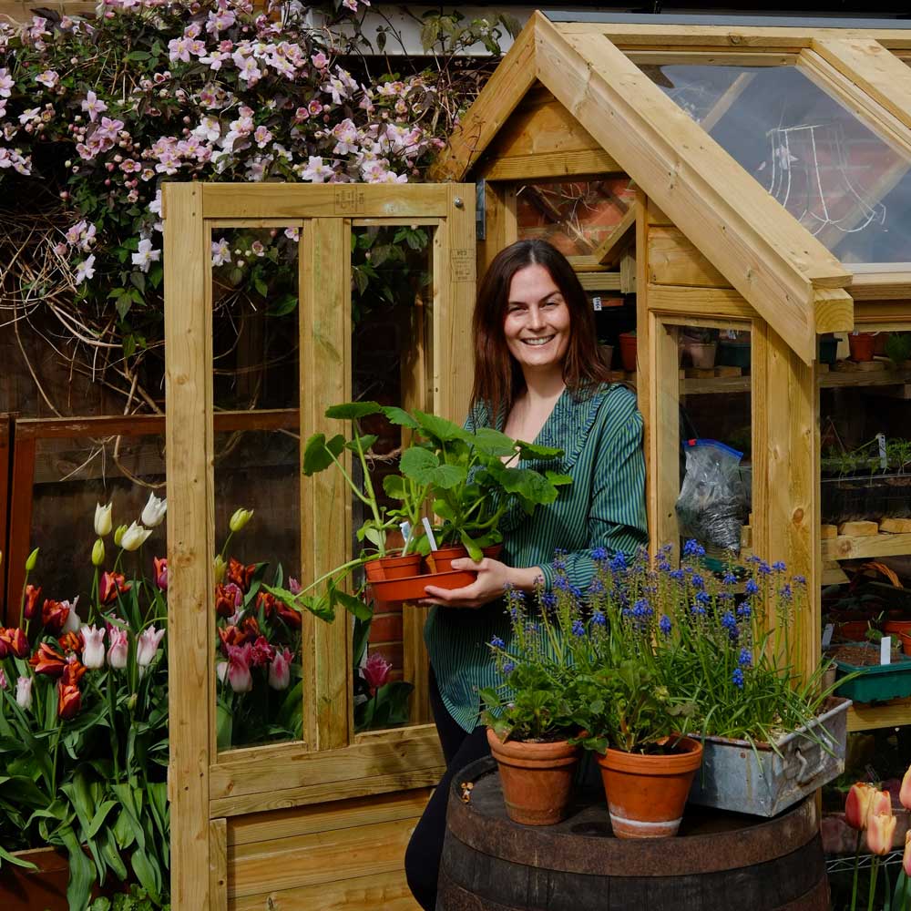 Woman holding potted plants in front of a wooden greenhouse with flowers and plants around.