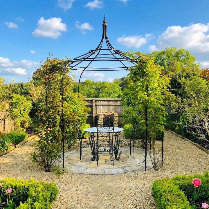 Agriframes Gothic Gazebo on a circular stone patio with plants growing up the frame and a table and chairs set underneath. 