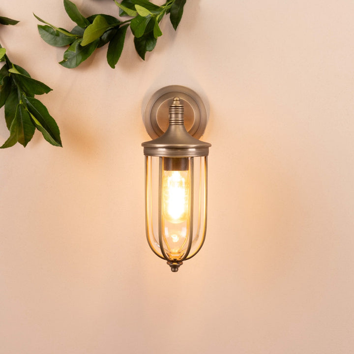 Front on image of the Agriframes Hampstead Outdoor Wall Lamp in a Brass finish with its bulb switched on, mounted on a neutral background.