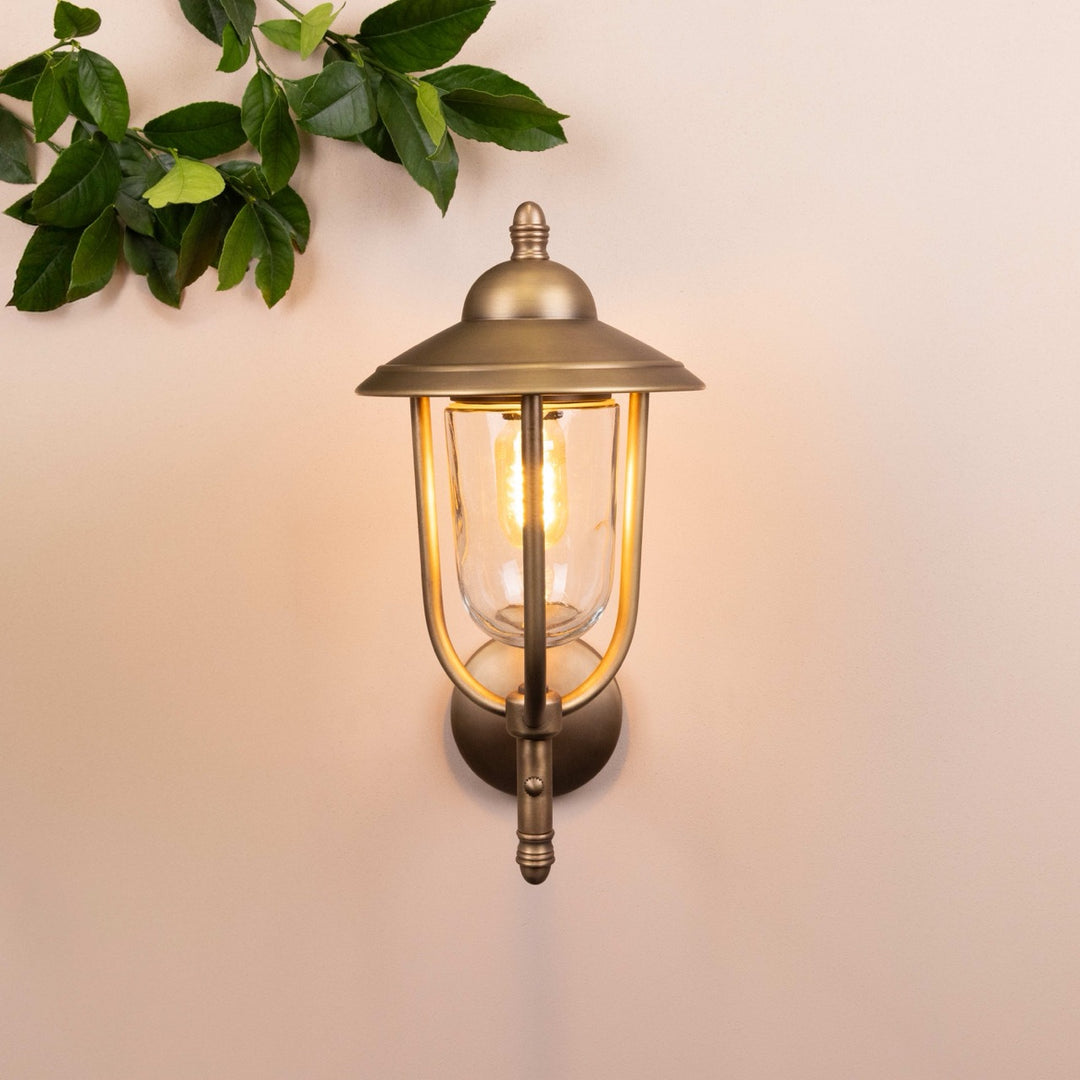 Front on image of the Agriframes Kensington Outdoor Wall Lamp in a Brass finish with its bulb illuminated, mounted on a neutral background. 