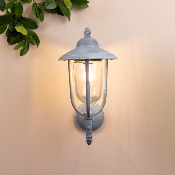 Front on image of the Agriframes Kensington Outdoor Wall Lamp in an Old Iron finish with its bulb illuminated, mounted on a neutral background. 