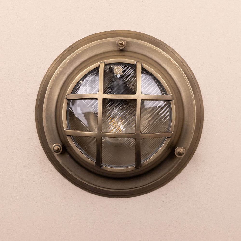 Front on image of the Agriframes Looe Bulk Head Round Lantern in a Brass finish with its bulb switched off, mounted on a neutral background. 