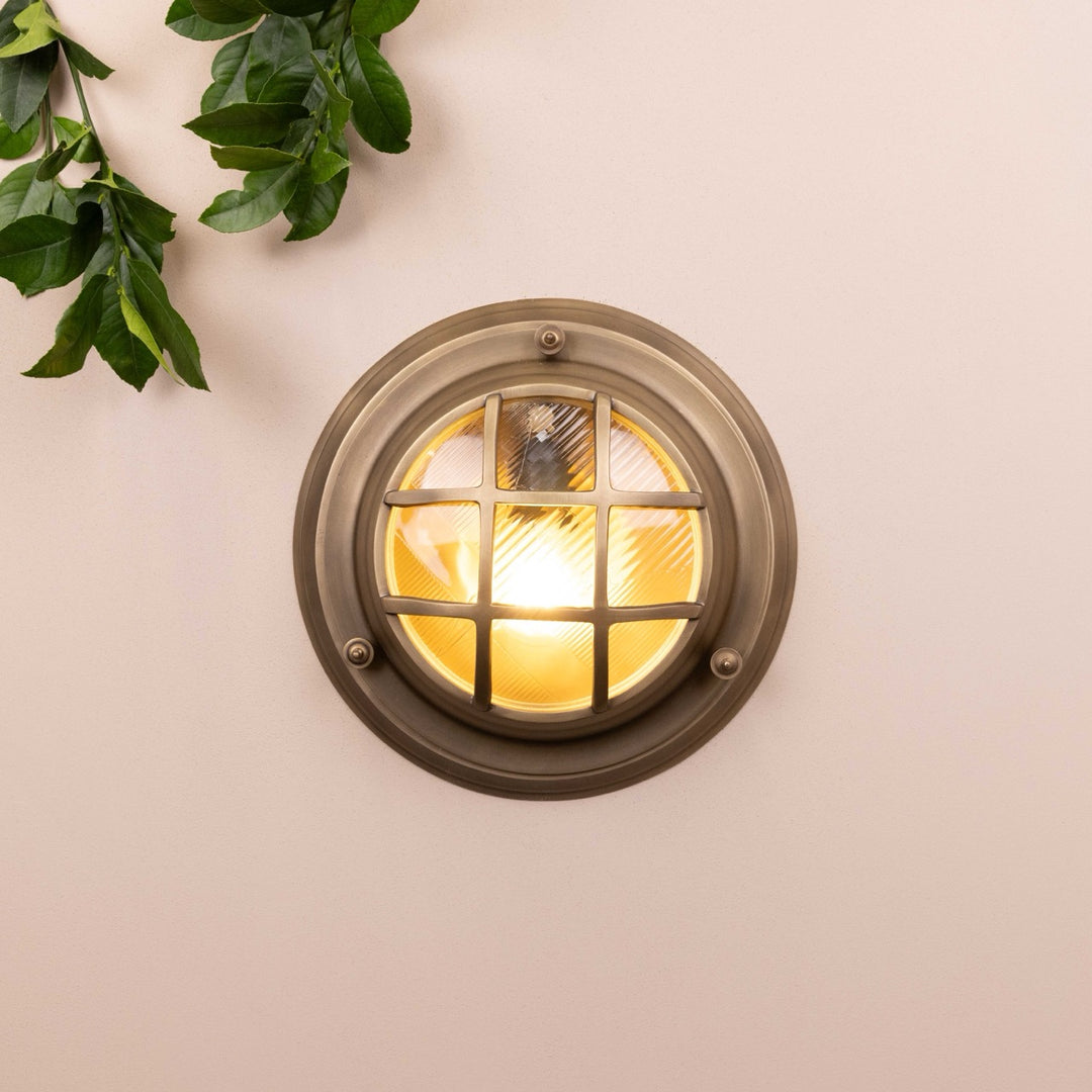 Front on image of the Agriframes Looe Bulk Head Round Lantern in a Brass finish with its bulb illuminated, mounted on a neutral background. 