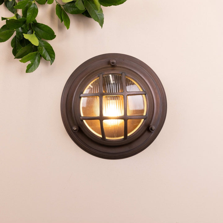 Front on image of the Agriframes Looe Bulk Head Round Lantern in a Bronze finish with its bulb illuminated, mounted on a neutral background. 