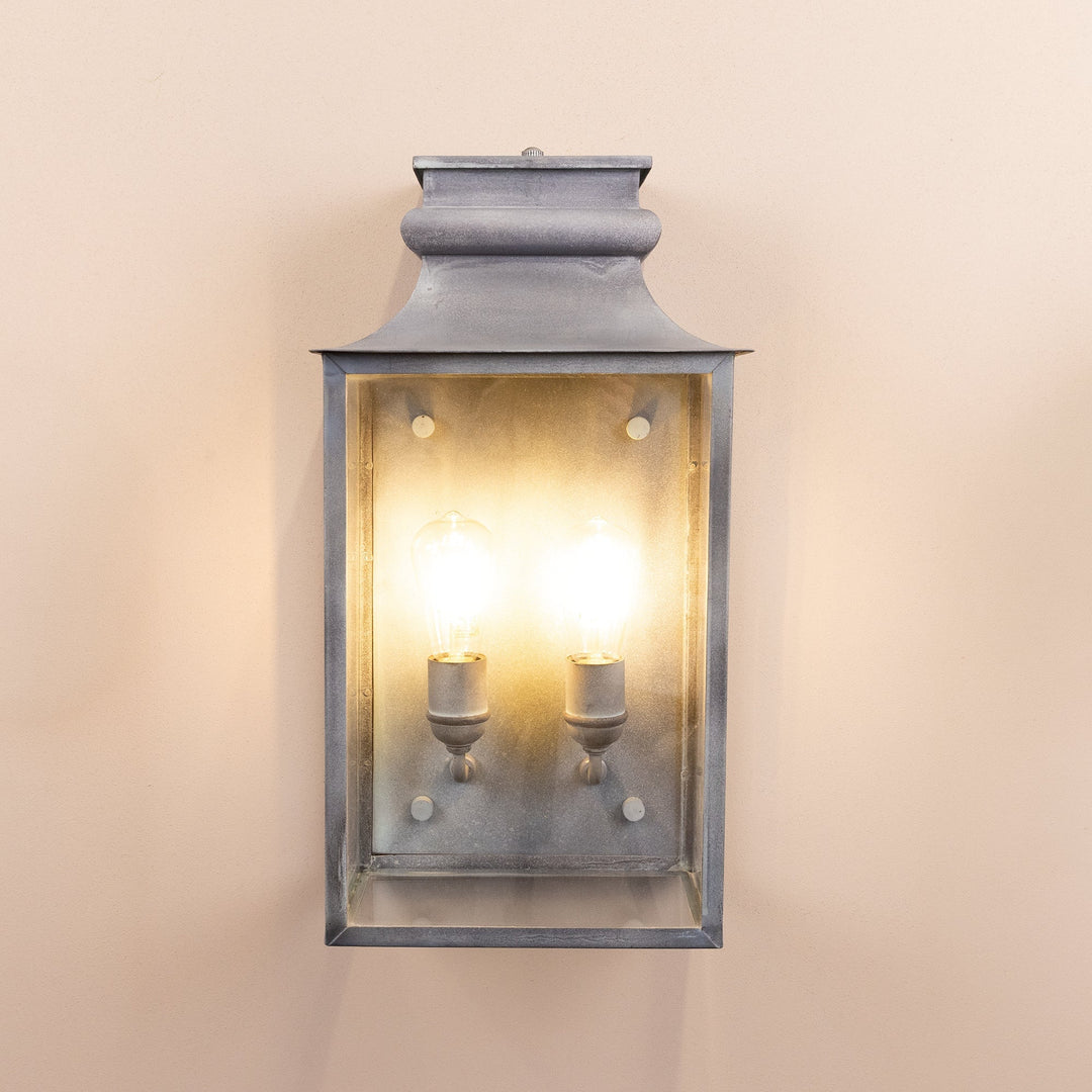 Agriframes Marlborough Coach Lantern in an Old Iron finish with its bulbs illuminated.  The lighting fixture is mounted on a neutral-coloured wall. 