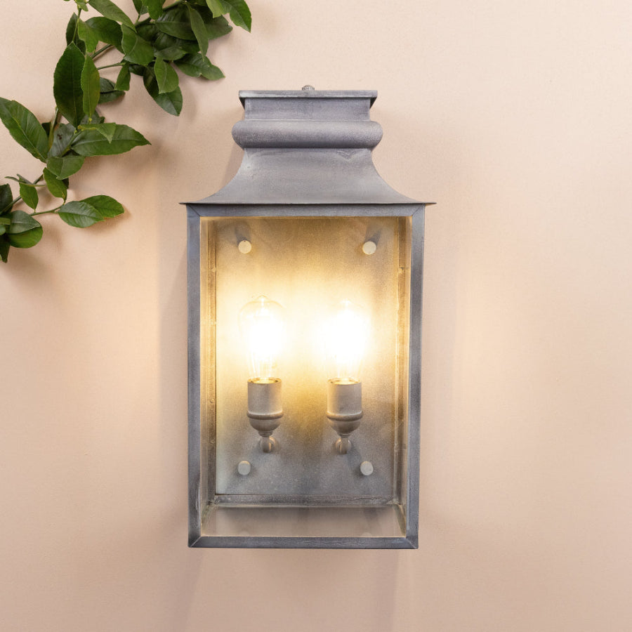 Agriframes Marlborough Coach Lantern with its bulbs illuminated mounted on a neutral background with a decorative plant next to it. 
