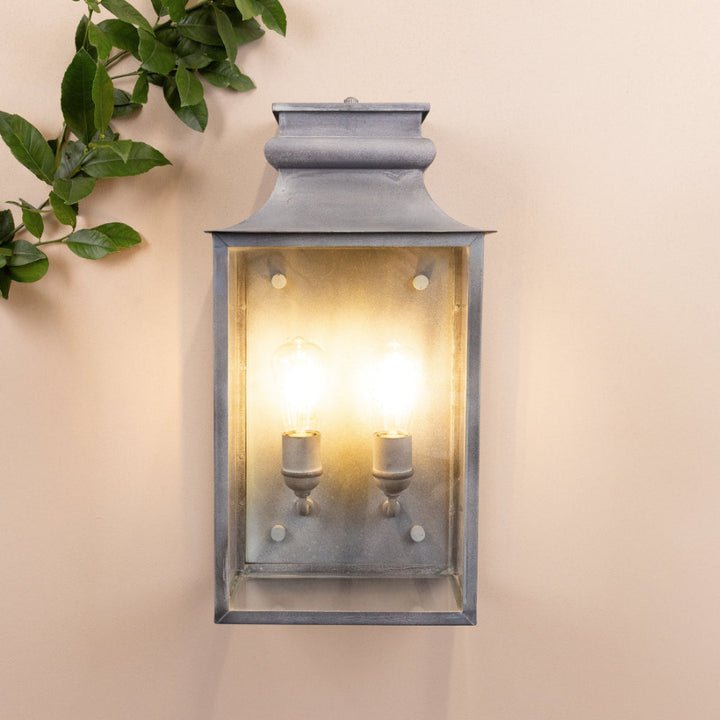 Agriframes Marlborough Coach Lantern with its bulbs illuminated mounted on a neutral background with a decorative plant next to it. 