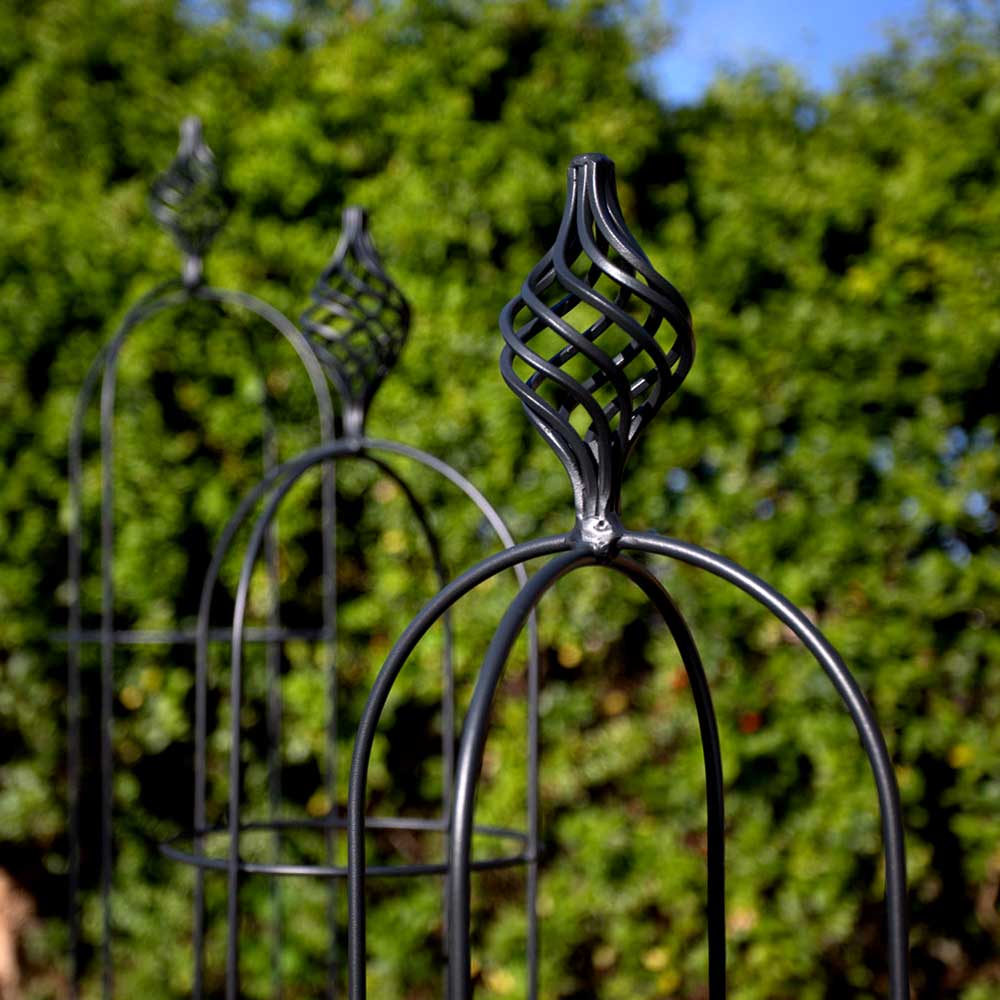 The top halves of three Agriframes matte black metal obelisks are positioned in a diagonal row in front of a hedgerow. 