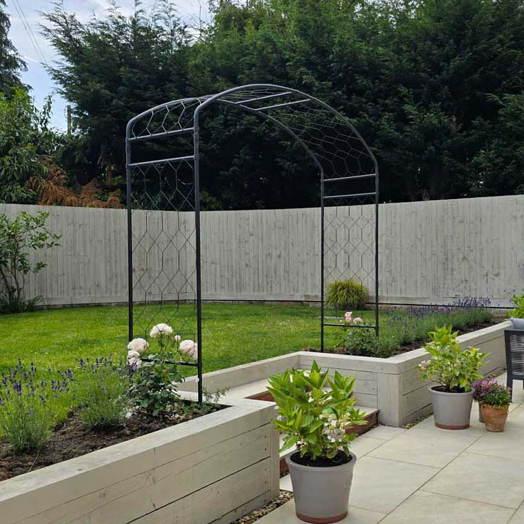 Garden scene with a metal arch, plants, and a paved area.