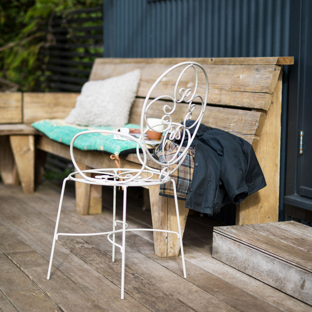 White Agriframes metal chair on a wooden deck with a wooden bench and cushions in the background.