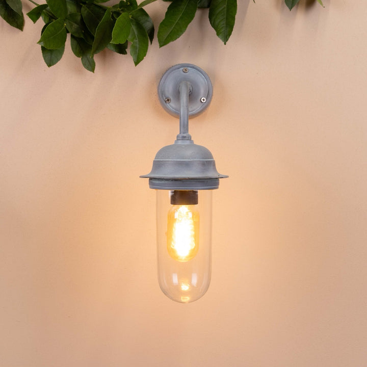 Front on image of the Agriframes Pewsey Outdoor Wall Lamp in an Old Iron finish with its bulb illuminated, mounted on a neutral background. 