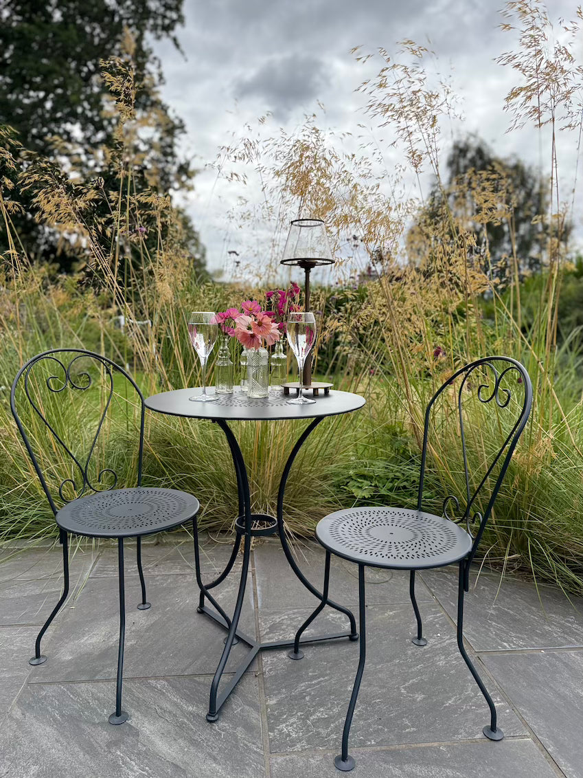 Provence 2 Seat Bistro Set on a patio with two glasses of wine, a vase of flowers and a tealight holder on the table. 