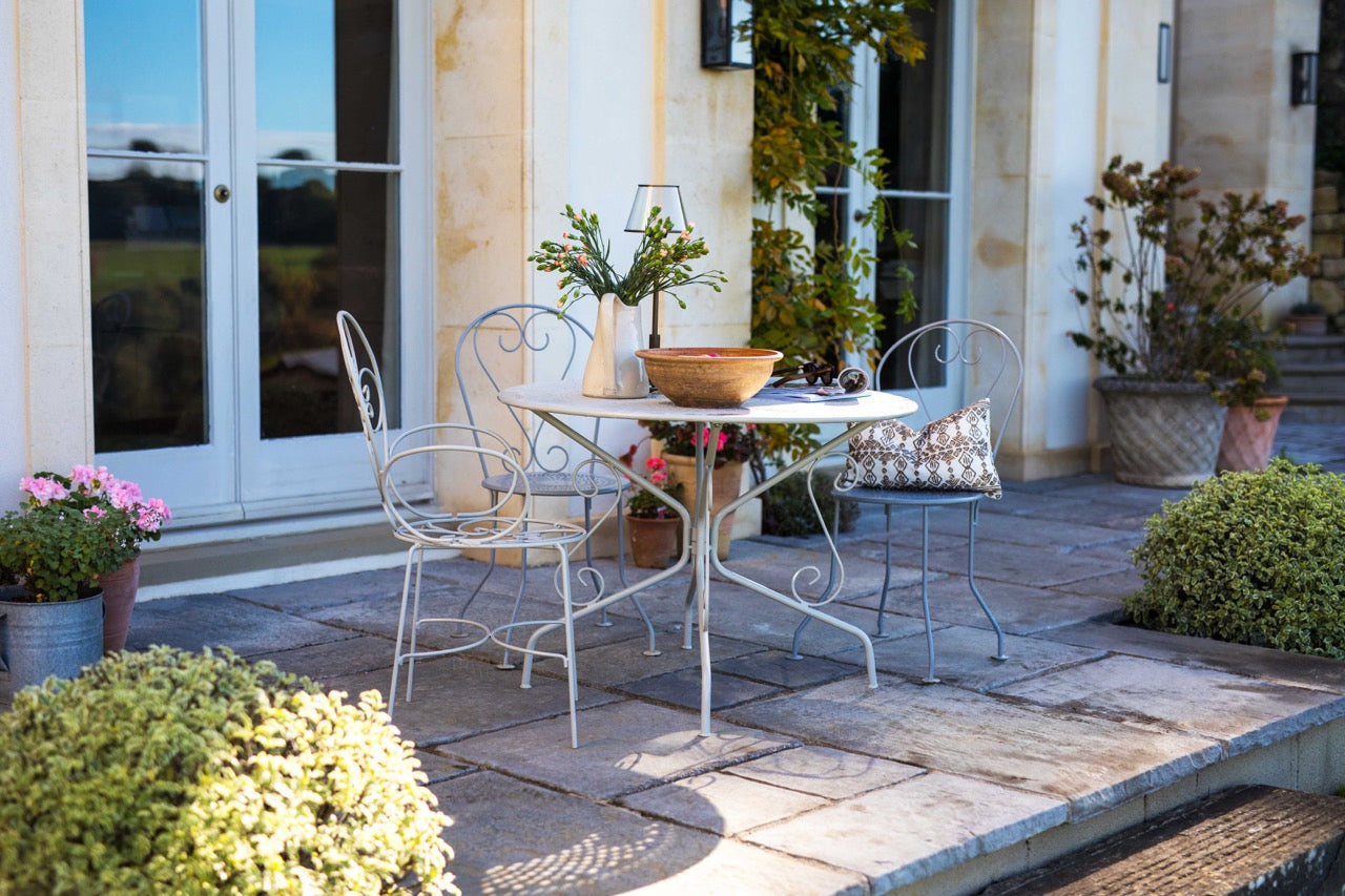Outdoor patio setting with a Agriframes Provence vintage 4 seater round table and chairs on a stone patio, surrounded by plants and flowers.