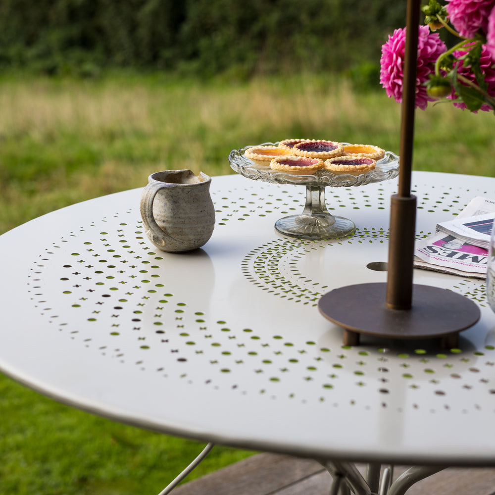 Round Agriframes Provence outdoor table with a mug, cake stand with pastries, and pink flowers in a garden setting.