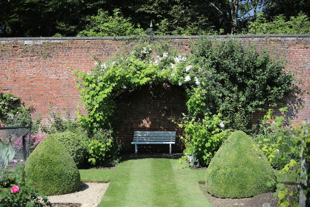 Agriframes round gazebo against a brick wall with a bench underneath and climbing roses covering the frame in a large walled garden.