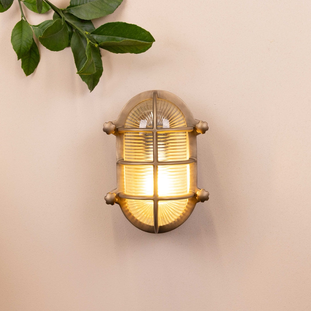 Brass oval-shaped light fixture mounted on a beige wall with decorative leaves in the top left corner. 
