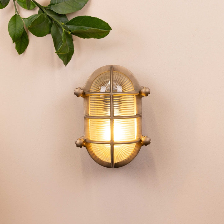 Brass oval-shaped light fixture mounted on a beige wall with decorative leaves in the top left corner. 
