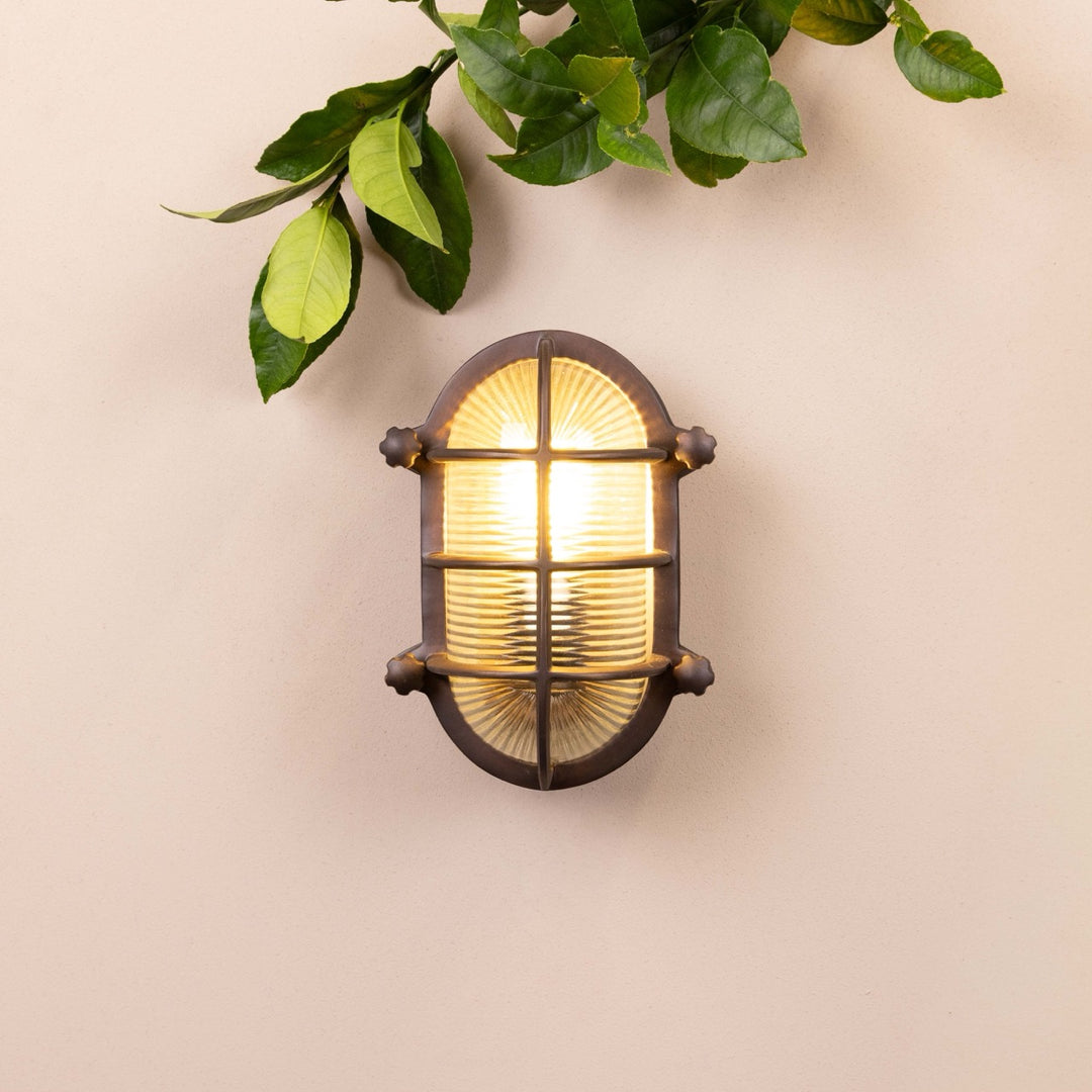 Bronze oval-shaped light fixture mounted on a beige wall with decorative leaves at the top of the frame. 