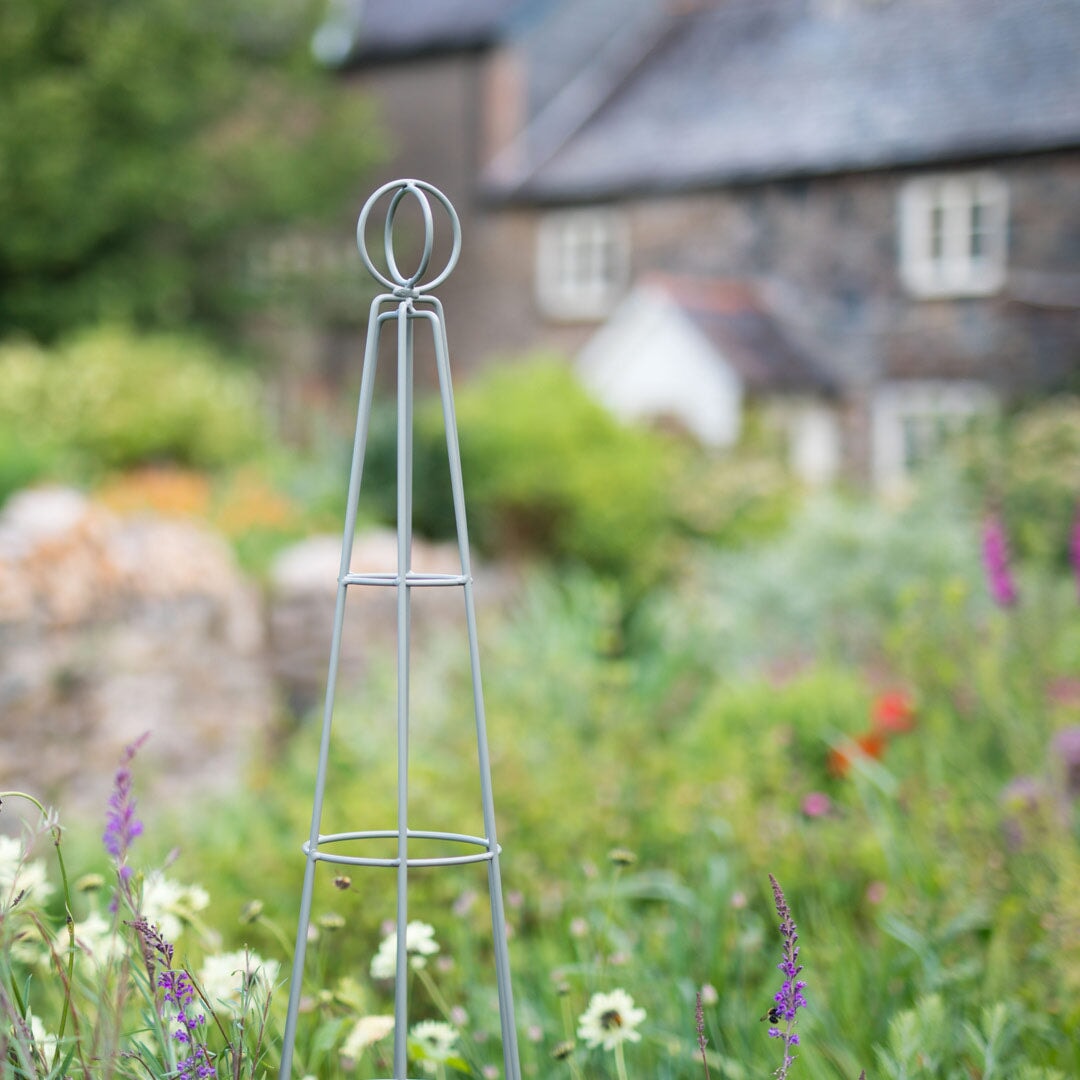 Agriframes Elegance Salisbury obelisk in a sage green finish in a flowerbed with a cottage in the background. 