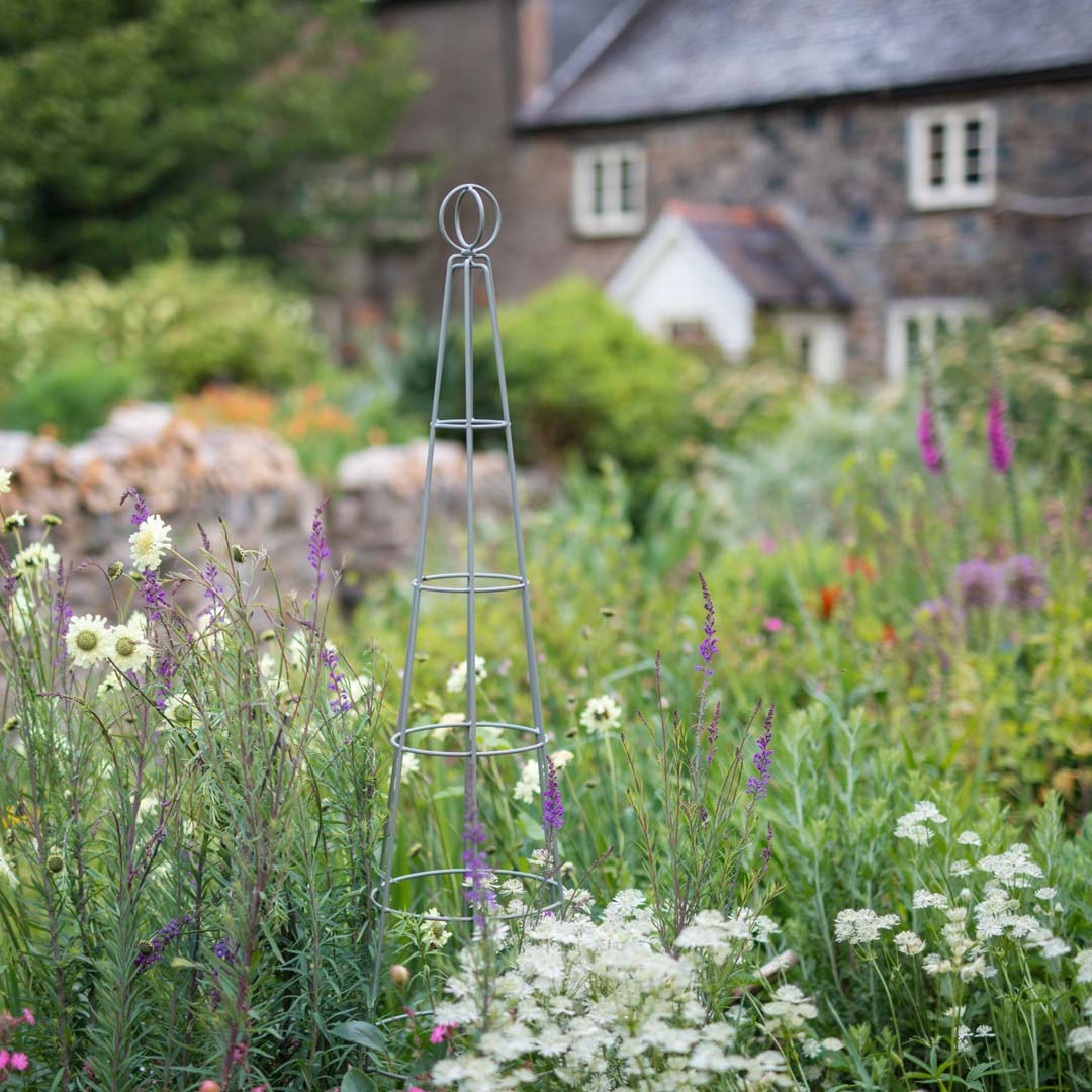 Agriframes Elegance Salisbury Obelisk in a sage green finish in a flowerbed with a cotage in the background.