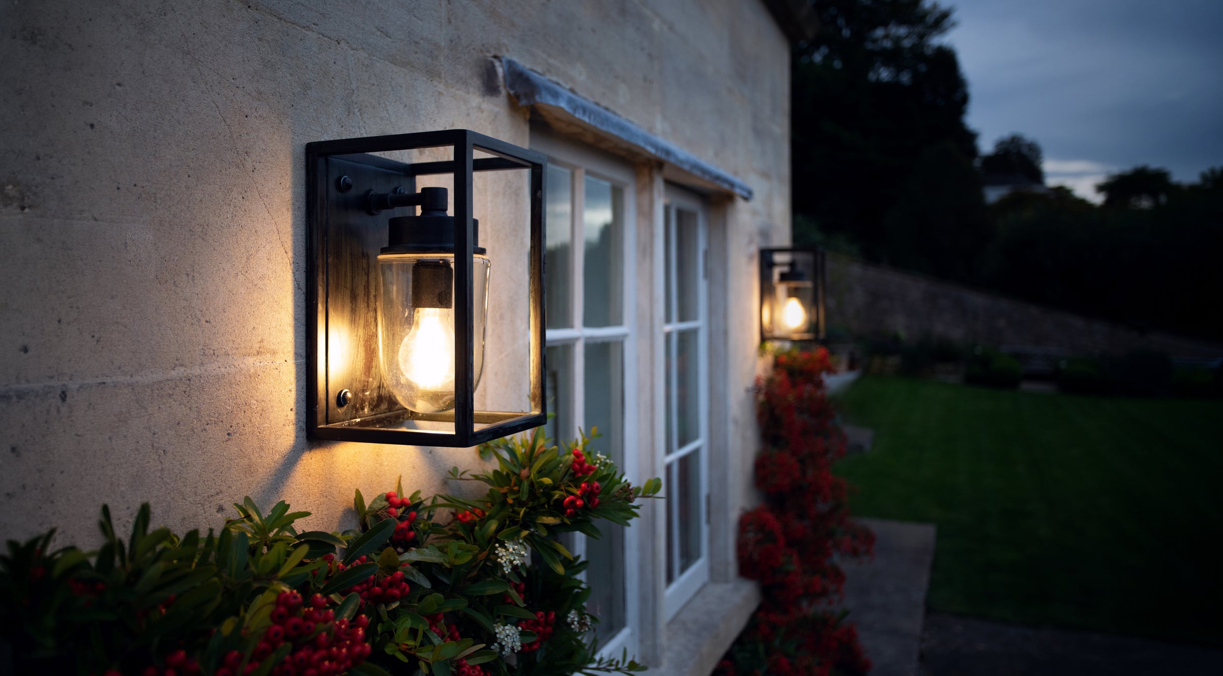 Two agriframes sherbourne outdoor open box lanterns installed on a stone house exterior either side of a window with plants growing around the lights. 