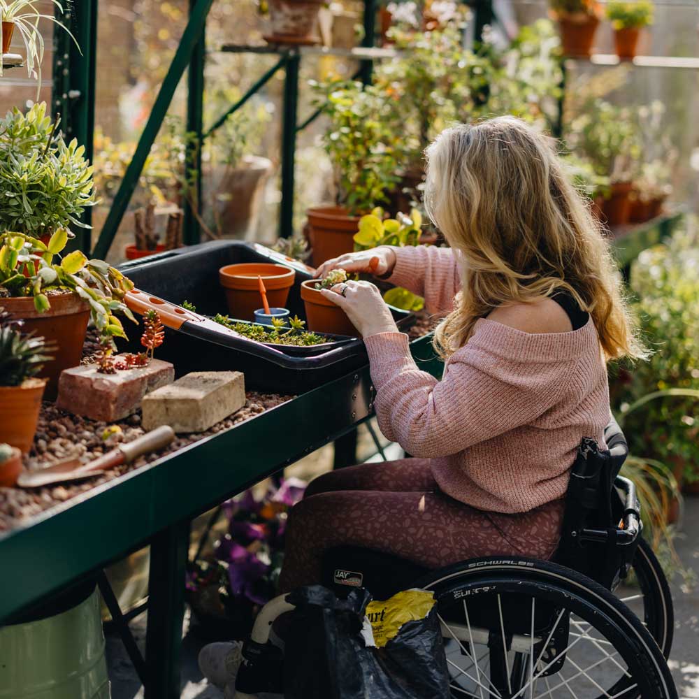 A wheelchair-bound woman gardening in a busy greenhouse.