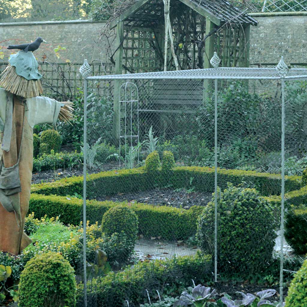 Tall Agriframes Elegance Vegetable Cage with sprial finials installed over a vegetable patch next to a scarecrow in a large garden